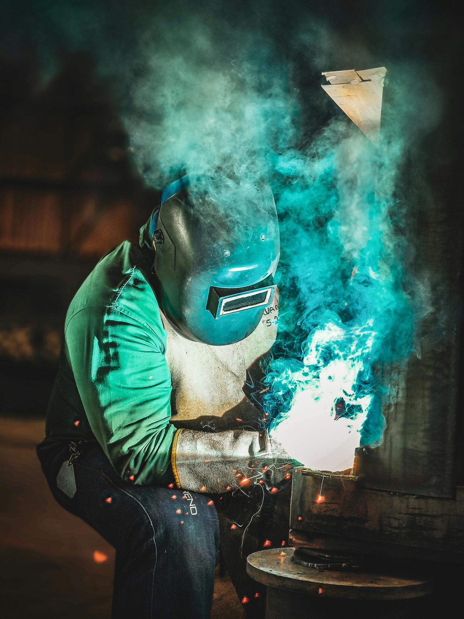 Welder wearing a protective helmet, gloves, and a green shirt works on a metal piece with intense blue flames and sparks, surrounded by smoke in a dimly lit industrial setting.