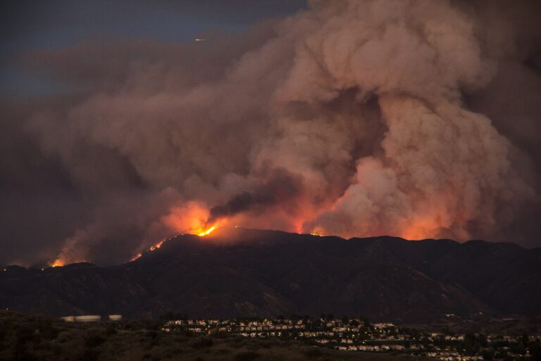 Wildfire burning across Santa Clarita California hills at night with heavy smoke rising above residential neighborhood in foreground.