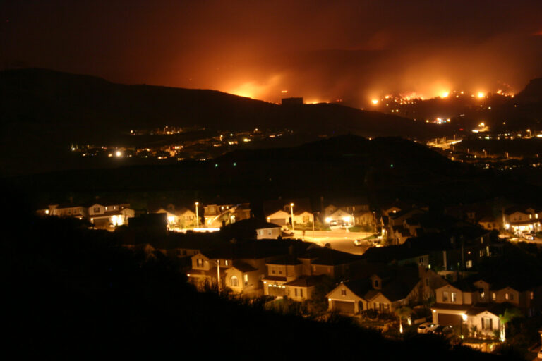 Wildfire burning on hillsides at night near a residential neighborhood in Santa Clarita, California, with flames glowing across the ridgeline behind rows of homes.