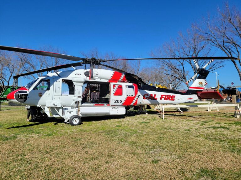 A photograph of a red and white Cal Fire helicopter on an open grass park with trees behind it.