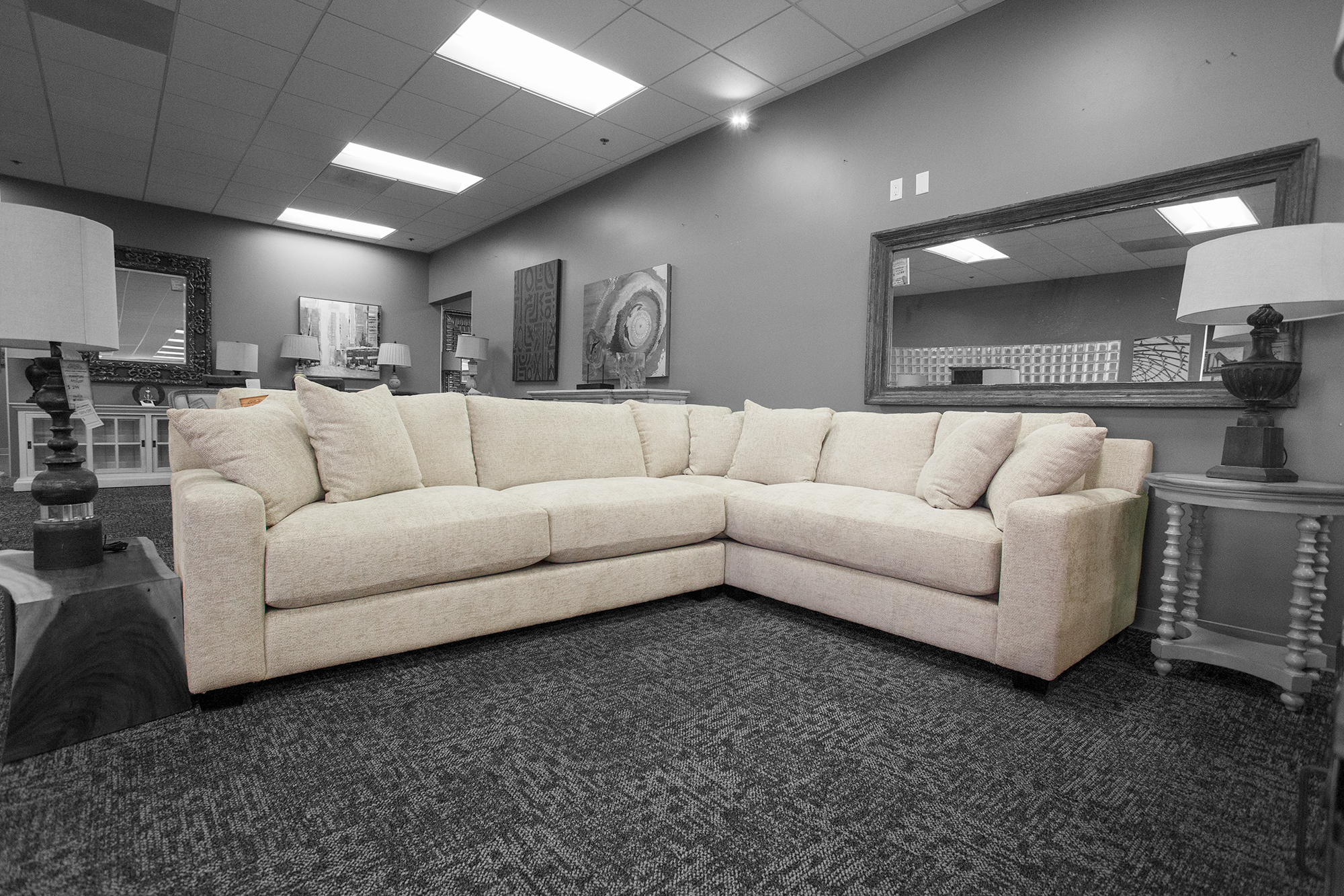 Cream-colored L-shaped sectional sofa with plush cushions, displayed in a showroom with gray walls, large wall mirrors, and accent lamps on each side. The sofa sits on a dark textured carpet and features a modern, minimalist design.