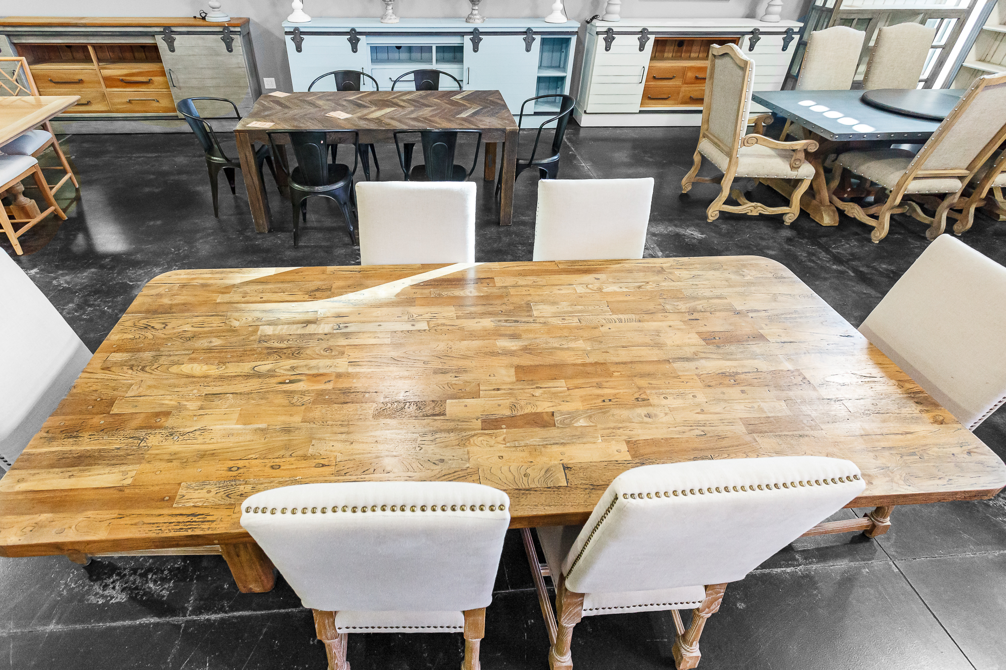 Oversized reclaimed wood dining table with nailhead-trimmed white chairs, surrounded by additional dining sets.
