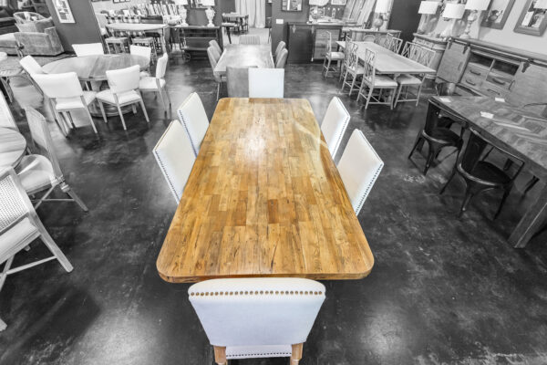 Long rectangular dining table made from reclaimed floorboards, surrounded by cream-colored nailhead chairs in a furniture showroom.