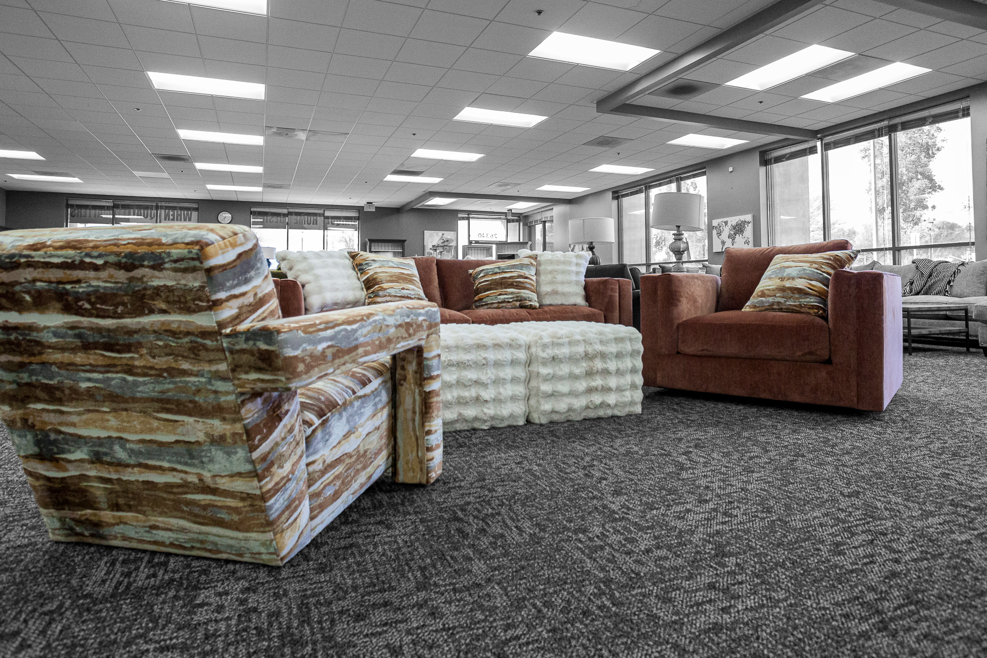 A living room setup featuring modern furniture, including a wide armed vogue style chair, two puffy white ottomans, and a red sofa and swivel chair. 