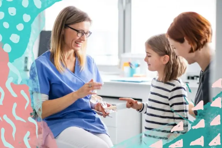A smiling dentist showing a set of dentures to a mother and her young daughter.